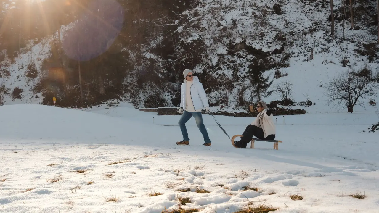 Paar beim Rodeln im verschneiten Gelände nahe dem Sport- und Wellnesshotel Held****s. Winteraktivität und Sonnenschein.