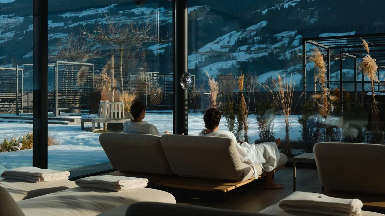 Gäste entspannen in Bademänteln auf Liegen im Ruheraum des Sport- und Wellnesshotel Held****s mit Blick auf verschneite Berge.
