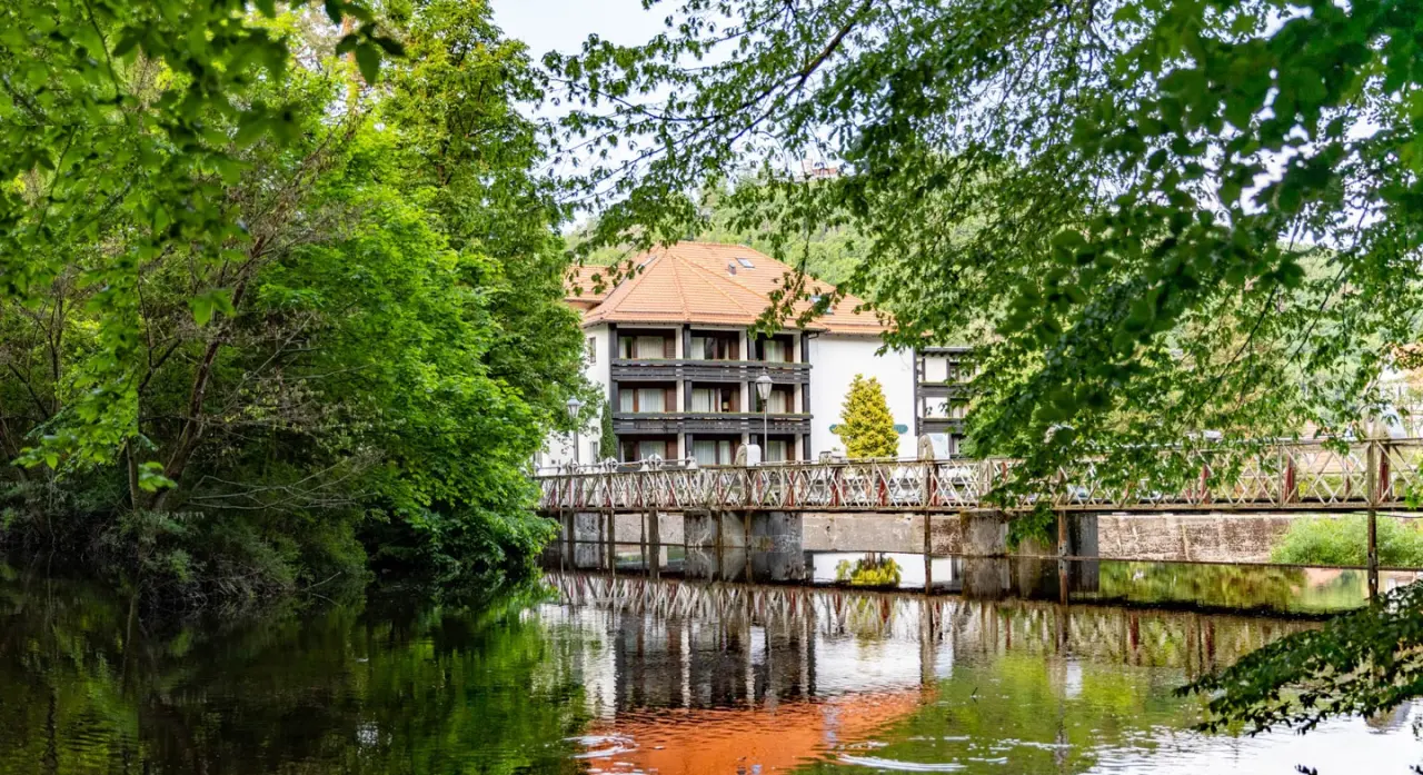 Mühl Vital Resort von einem Fluss aus gesehen, umgeben von üppigem Grün und einer historischen Brücke.