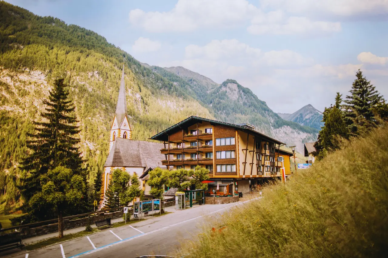 Aussenansicht der Nationalpark Lodge Grossglockner mit traditioneller Architektur, umgeben von Bergen und einer Kirche.