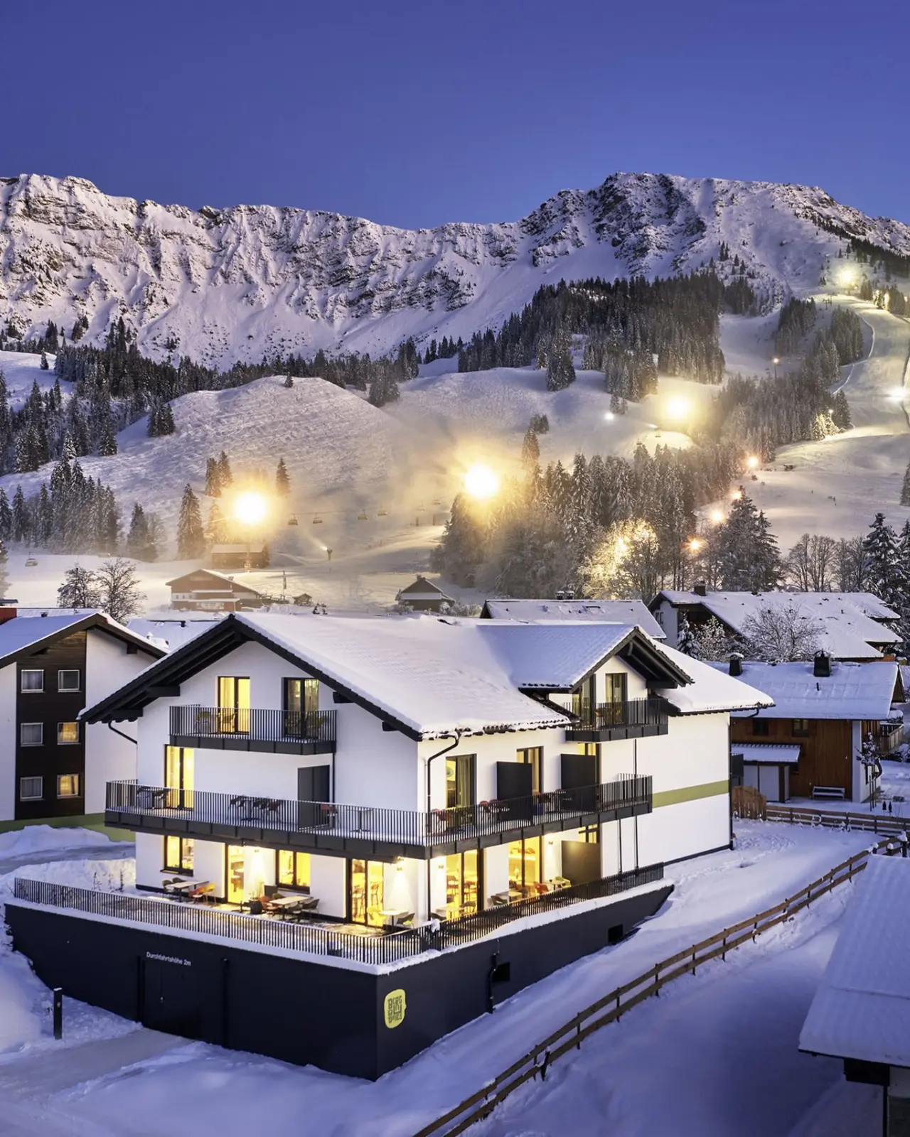 Winterliches Parkhotel Jordanbad bei Nacht. Beleuchtetes Gebäude vor verschneiten Alpen und beleuchteten Skipisten.