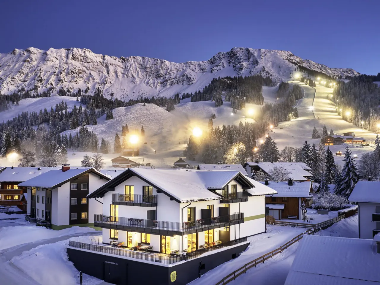 Winterliche Aussenansicht des Parkhotel Jordanbad bei Dämmerung, mit beleuchteten Skipisten und verschneiten Bergen.