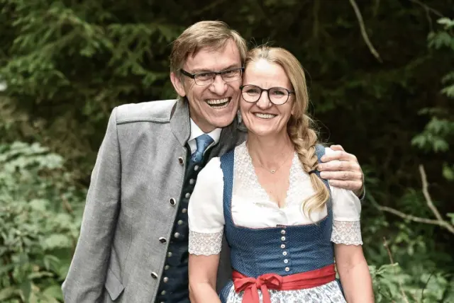 Freundliches Gastgeberpaar des ALMGUT Mountain Wellness Hotels in traditioneller Tracht vor grüner Naturkulisse.