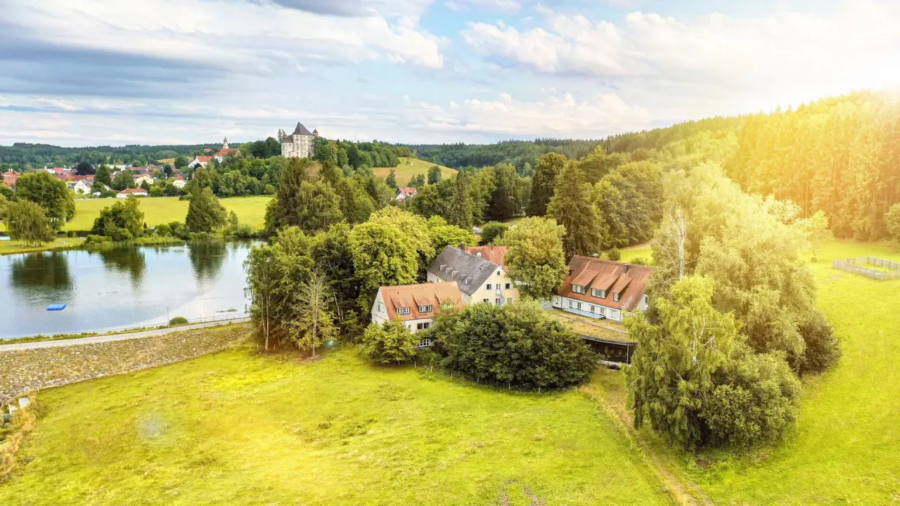 Luftaufnahme des Bad Clevers Gesundheitsresort & Spa mit Hotelgebäuden, See, Wald und Schloss im Hintergrund bei Sonnenschein.