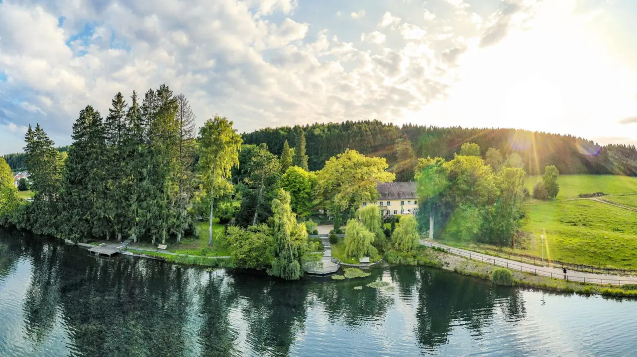Panoramablick auf das Bad Clevers Gesundheitsresort & Spa am See, umgeben von grüner Natur und Wald bei Sonnenuntergang.