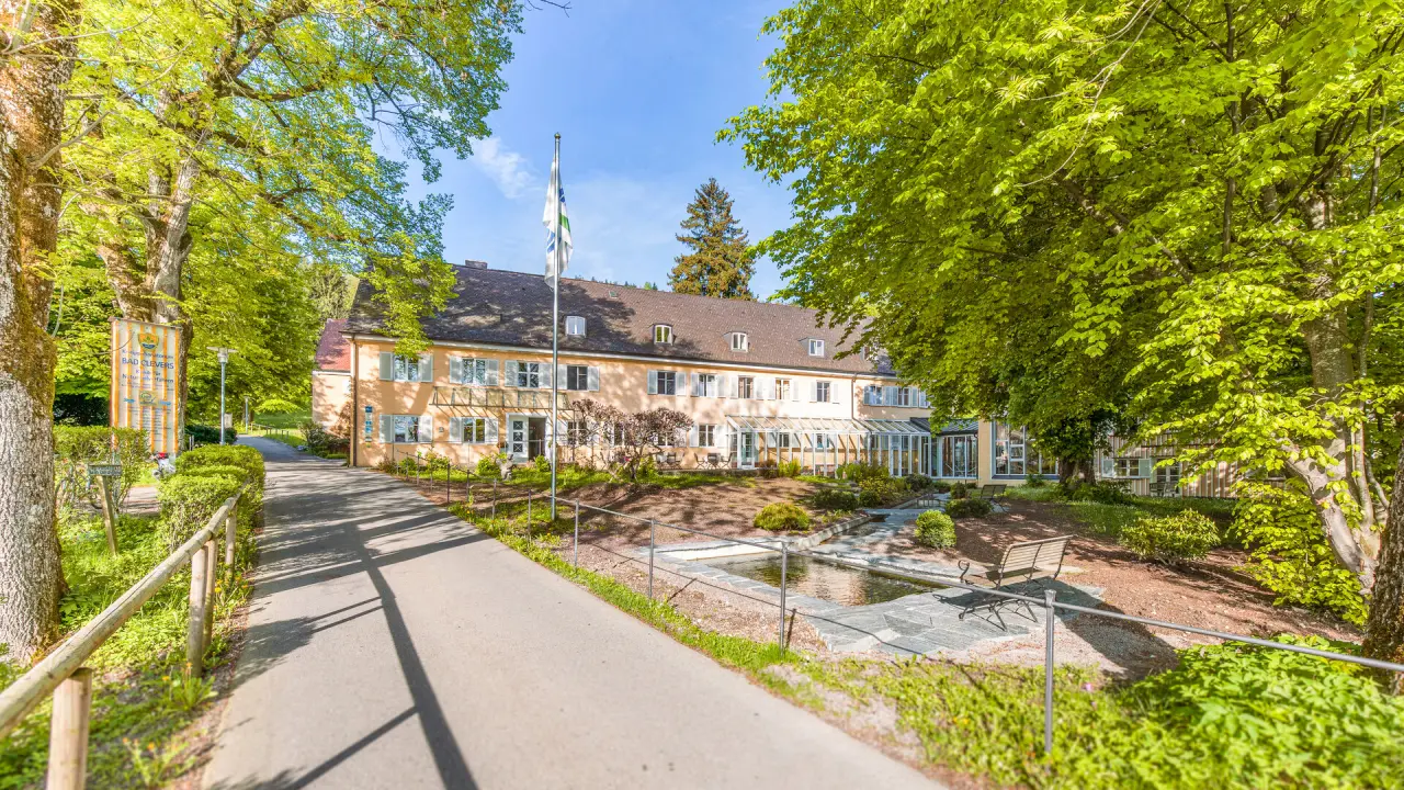 Aussenansicht des Bad Clevers Gesundheitsresort & Spa mit grünen Bäumen und sonnigem Weg zum Haupteingang.