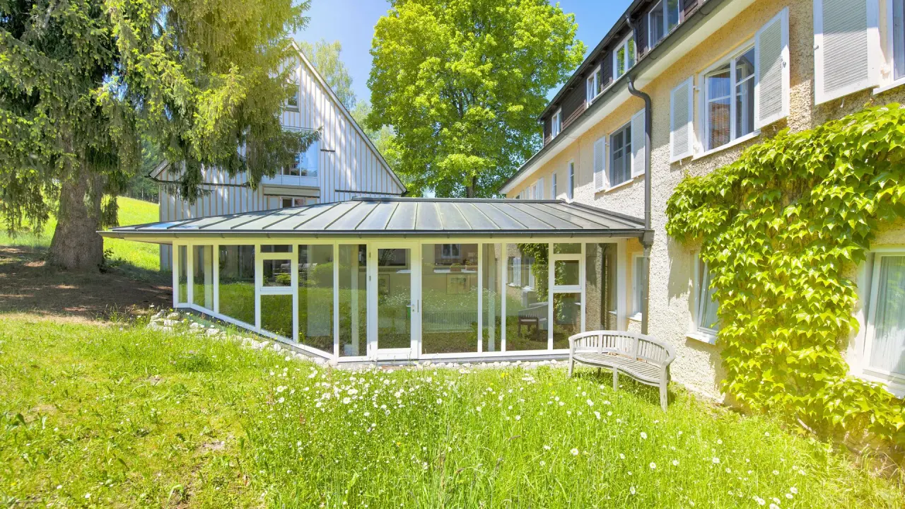 Grüne Aussenansicht des Bad Clevers Gesundheitsresort & Spa mit Wintergarten, blühender Wiese und alten Bäumen an einem sonnigen Tag.