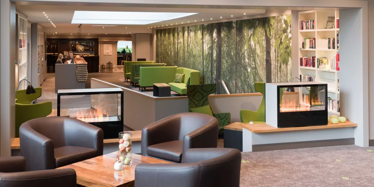 Einladende Hotellobby mit Kamin, grünen Sitzbereichen und Waldmotiv im Hotel Munte am Stadtwald.