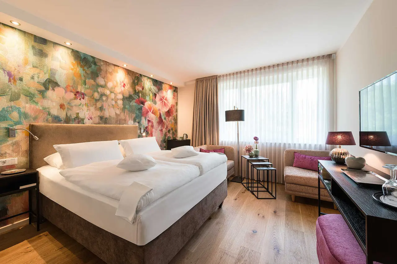 Geräumiges Hotelzimmer im Hotel Munte am Stadtwald mit Doppelbett, floraler Wandgestaltung und gemütlicher Sitzecke.