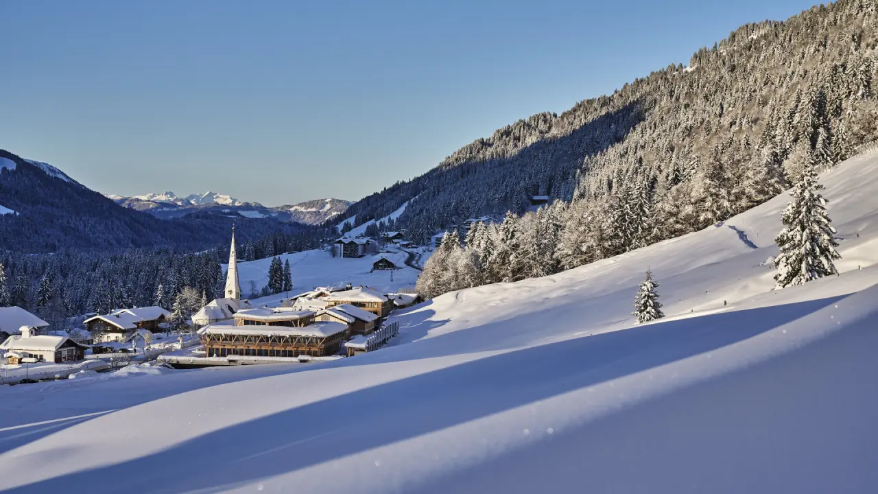 Winterlandschaft mit dem Hubertus Mountain Refugio Allgäu und verschneiten Bergen unter blauem Himmel.