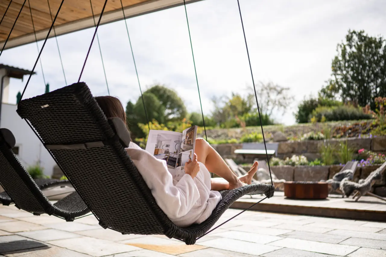 Gast im Bademantel entspannt in einer Hängeliege im Gartenbereich des Hotel Johanneshof, umgeben von Grün und Steinmauern.