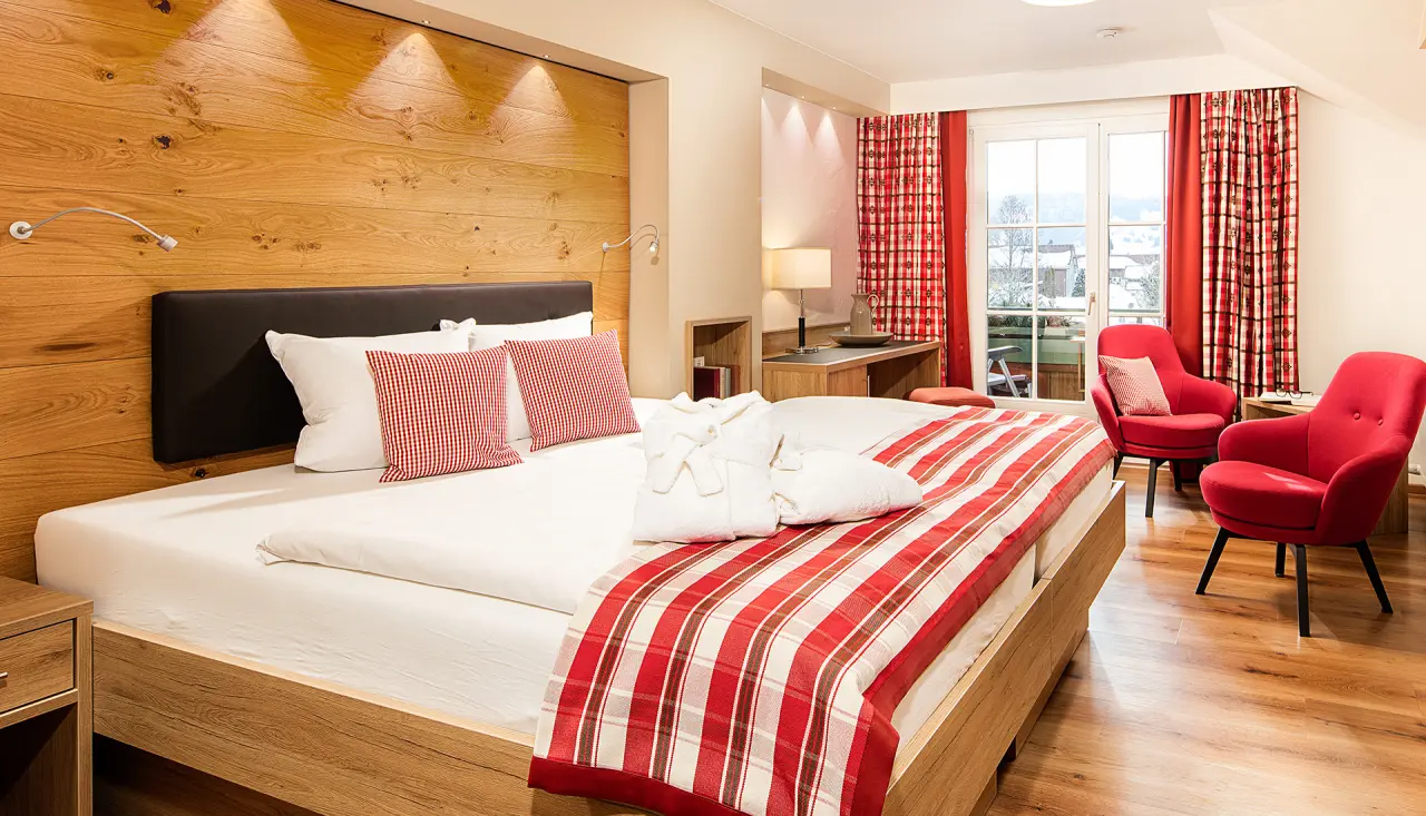 Gemütliches Doppelzimmer im Hotel Johanneshof mit Holzwand, roter Bettdecke, Bademänteln und Sitzbereich mit Balkonblick.