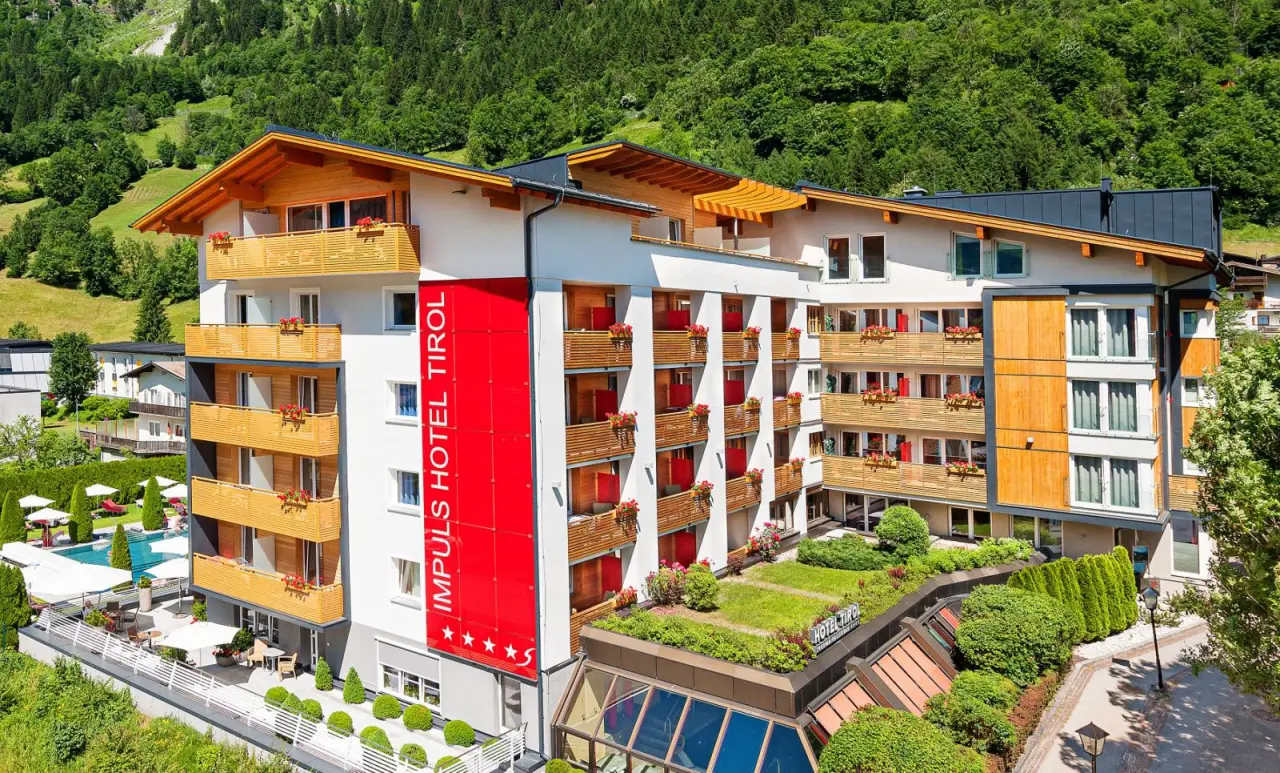 Aussenansicht des Impuls Hotel Tirol mit modernen Balkonen, rotem Banner und Aussenpool vor grüner Berglandschaft.