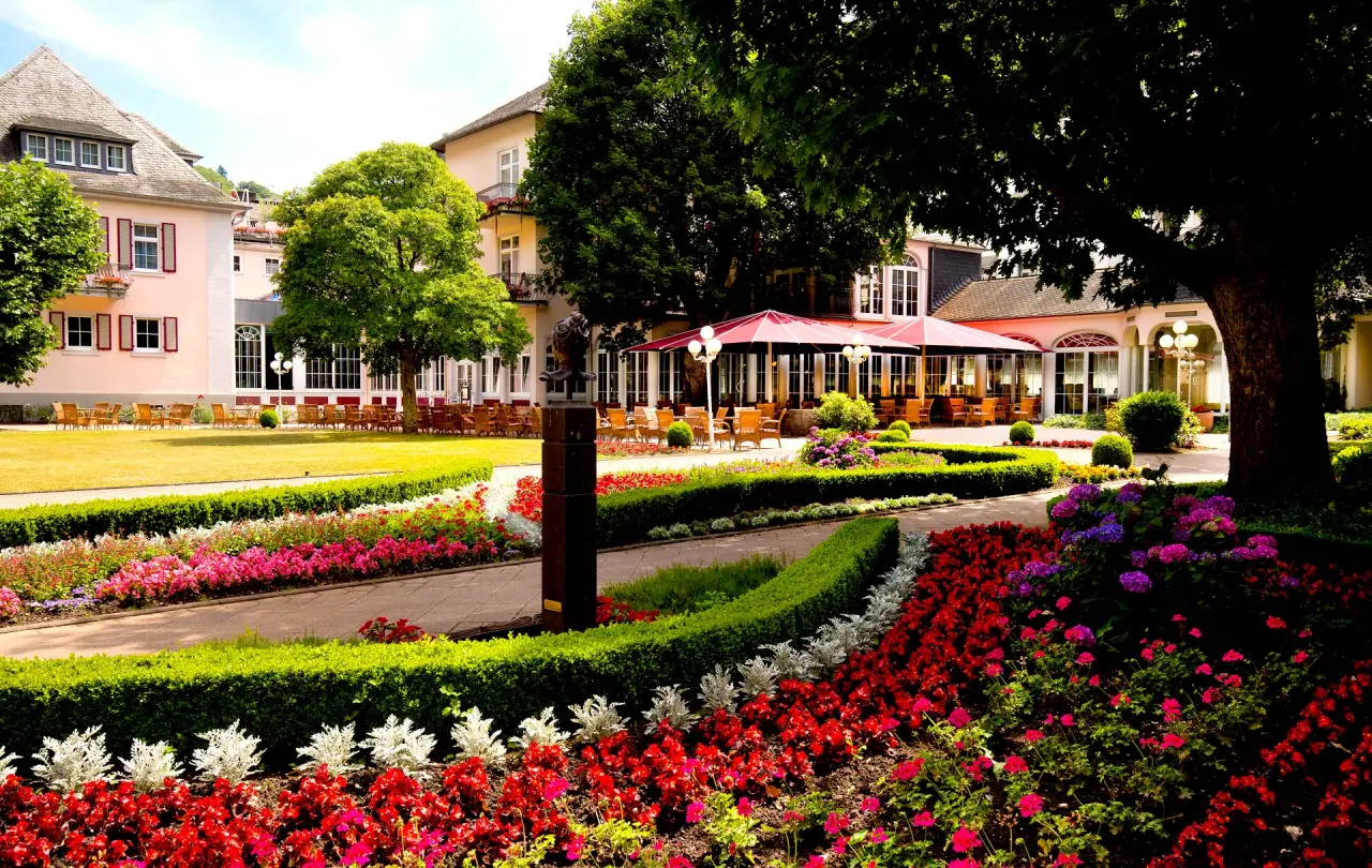 Aussenansicht von Häcker´s Fürstenhof Bad Bertrich mit gepflegtem Garten, bunten Blumen und sonniger Terrasse.