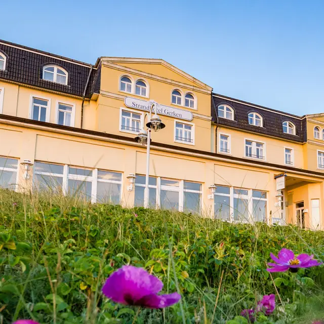 Aussenansicht des Strandhotel Gerken mit gelber Fassade und blühenden Pflanzen im Vordergrund unter blauem Himmel.