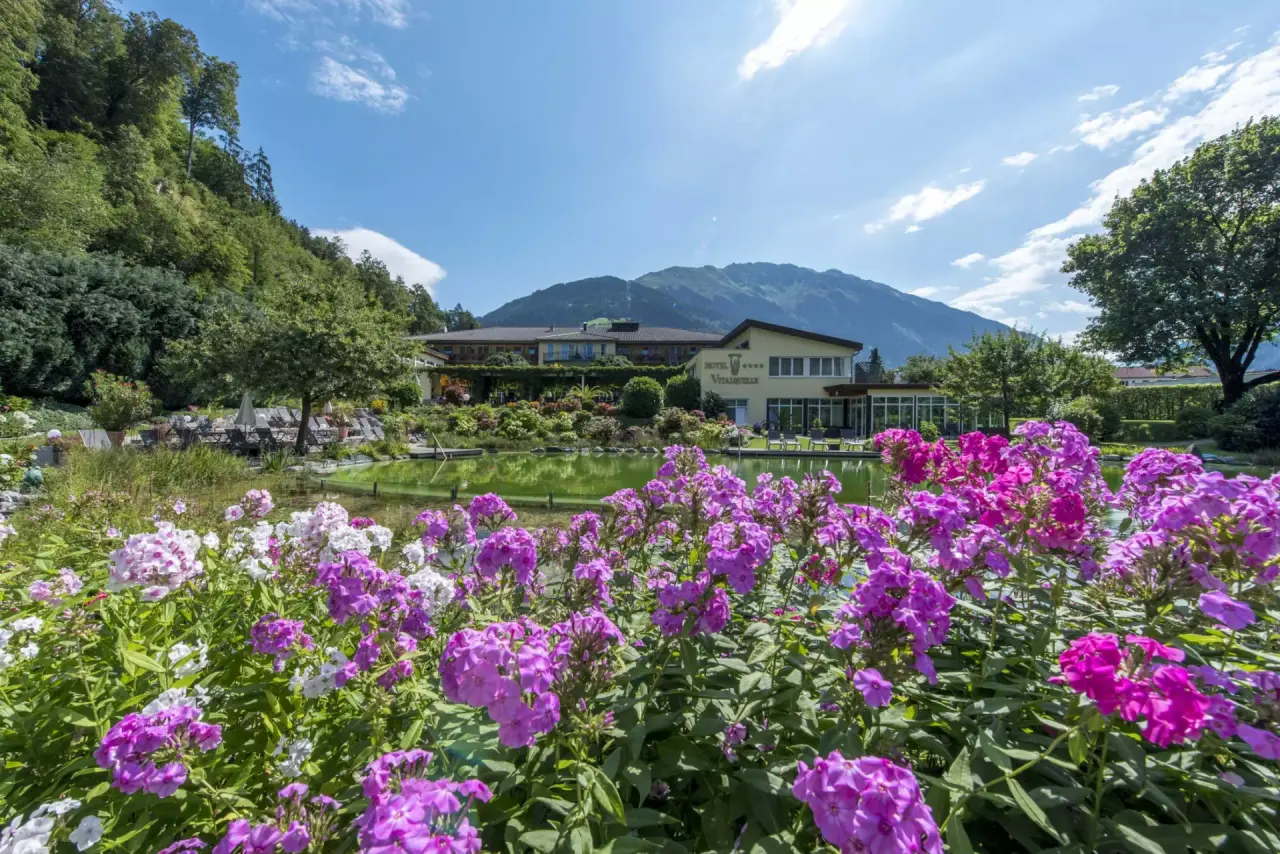 Aussenansicht des Hotel Vitalquelle Montafon mit blühendem Garten, Naturteich und Bergpanorama unter blauem Himmel.