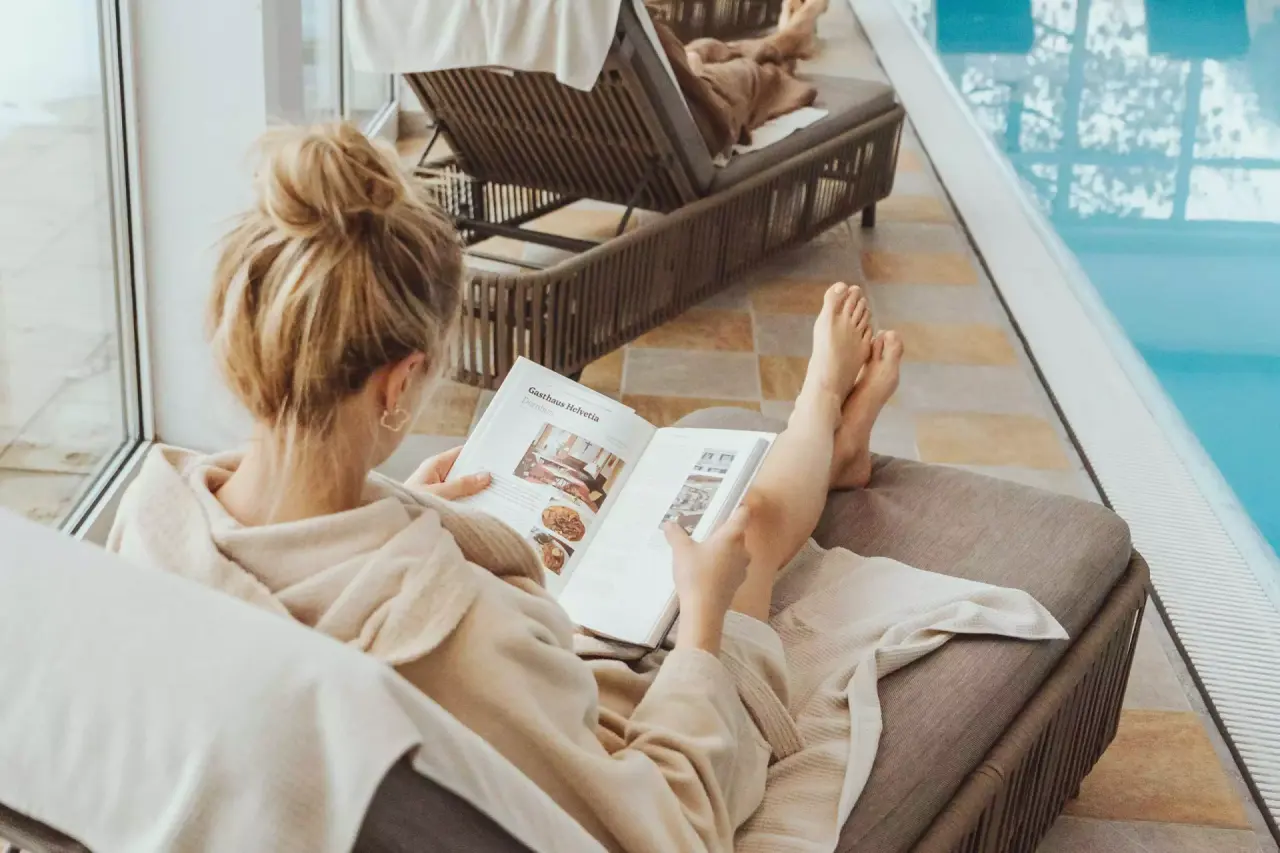 Gast entspannt im Bademantel auf einer Liege am hellen Innenpool der Vitalquelle Montafon, liest ein Magazin.
